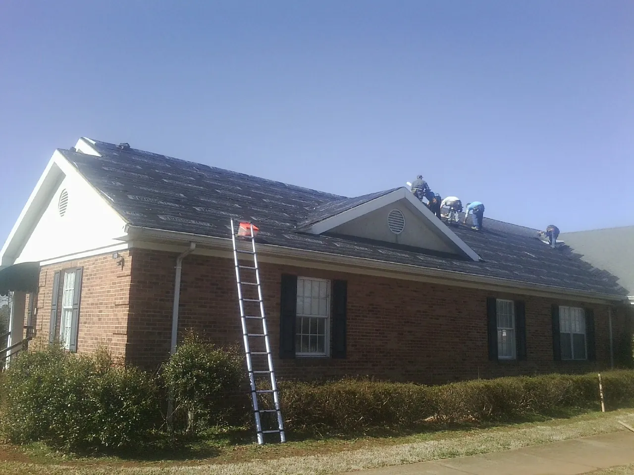 Crew tearing shingles off a brick ranch roof with a ladder set — Metal Roof Repair work by our crew in Winsted