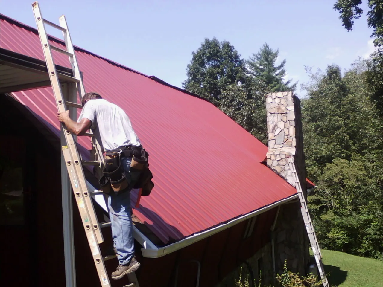 Roofer on a ladder beside a red metal roof and stone chimney — Soffit & Fascia Repair by Roofing Brothers in Winsted